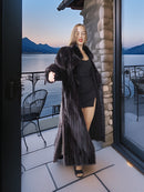 Woman in a long dark fur coat standing on a balcony with a scenic view of mountains and water.