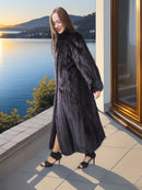 Woman in a long black fur coat standing by a window with a scenic view of water and mountains.