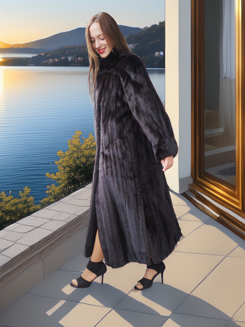 Woman in a long black fur coat standing by a window with a scenic view of water and mountains.