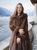 Woman wearing a brown fur coat by a lakeside with mountains in the background