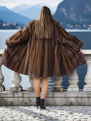 Person wearing a brown fur coat standing by a scenic lakeside with mountains in the background.