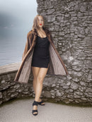 Woman in a long fur coat standing against a stone wall by a body of water.