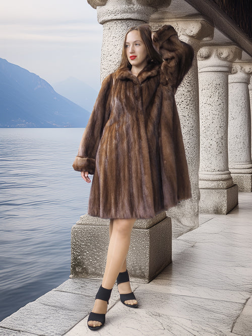 Woman in a fur coat standing by a lakeside with mountains in the background