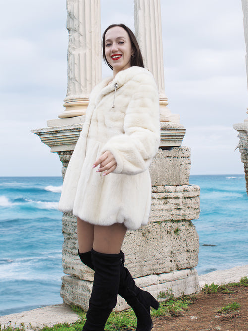 Beige Pearl Mink Fur Coat with hood, worn by a woman near ancient ruins by the sea. Features hook-eye closure, satin lining, and elegant warmth.