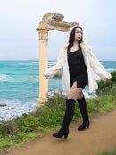 Elegant woman in a beige pearl mink fur coat with a hood, walking along a coastal path. The luxurious coat features a hook-eye closure and satin lining.
