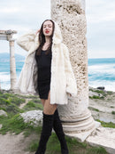 Beige Pearl Mink Fur Coat with hood, worn over a black dress near ancient ruins by the sea. Luxurious warmth with hook-eye closure and satin lining.