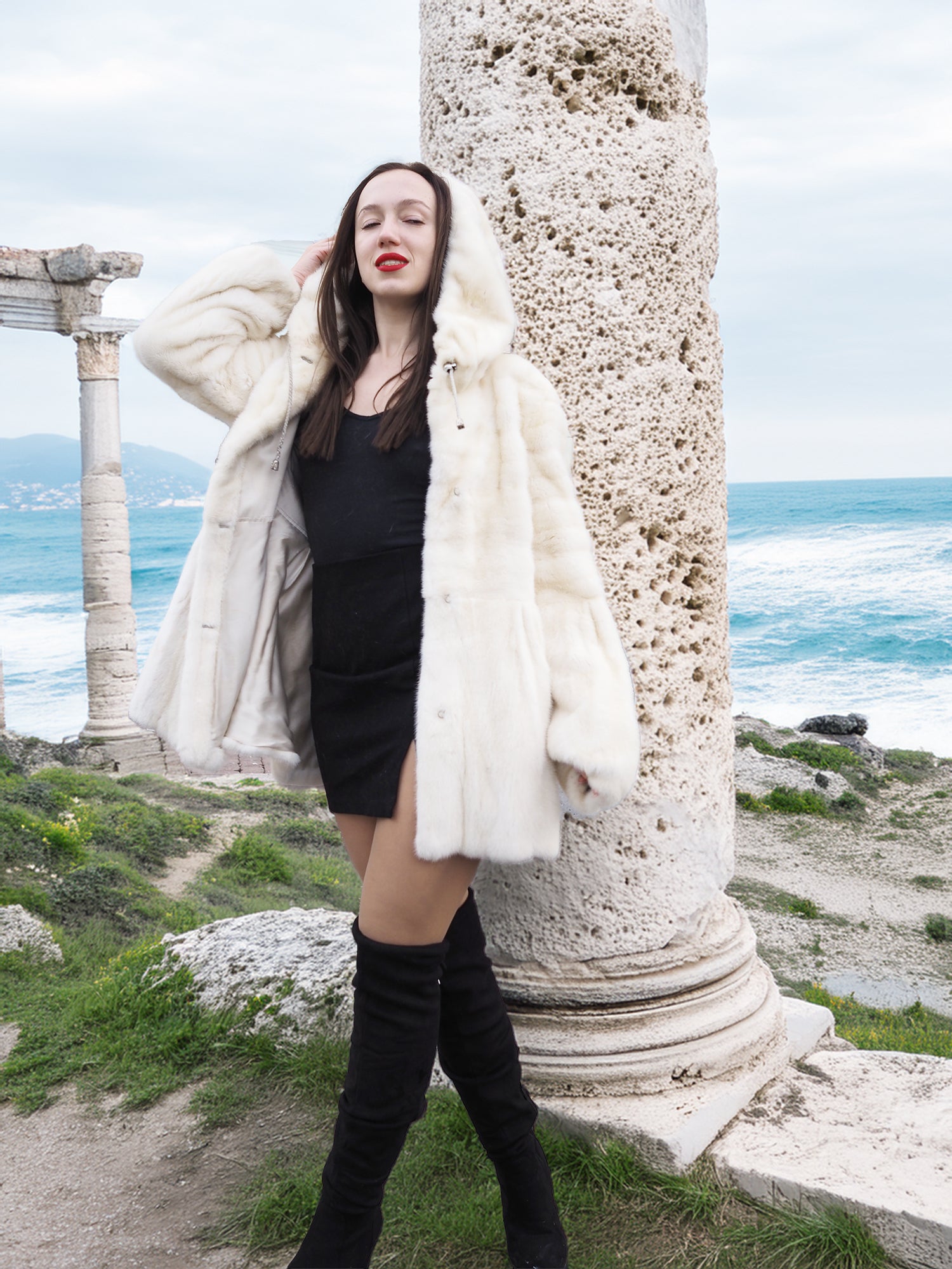 Beige Pearl Mink Fur Coat with hood, worn over a black dress near ancient ruins by the sea. Luxurious warmth with hook-eye closure and satin lining.