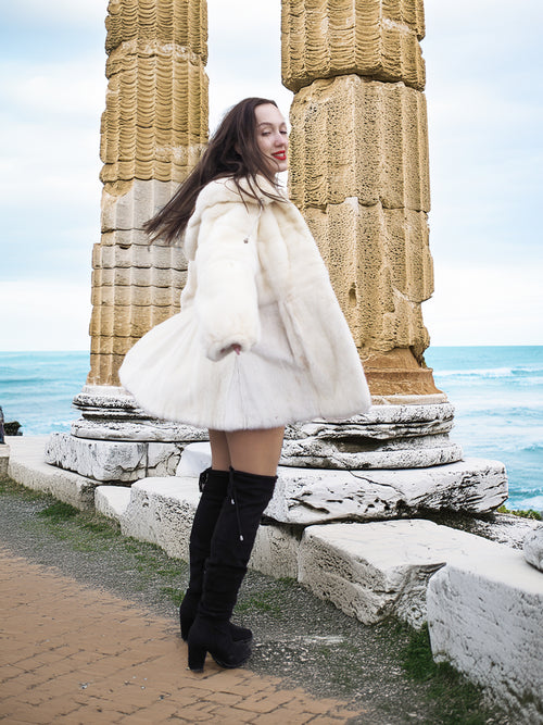 Elegant Beige Pearl Mink Fur Coat with hood, worn by a model near ancient ruins overlooking the sea. Luxurious warmth with hook-eye closure and satin lining.