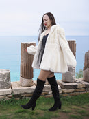Elegant Beige Pearl Mink Fur Coat with hood, worn by a model near ancient ruins overlooking the sea. Luxurious warmth with hook-eye closure and satin lining.