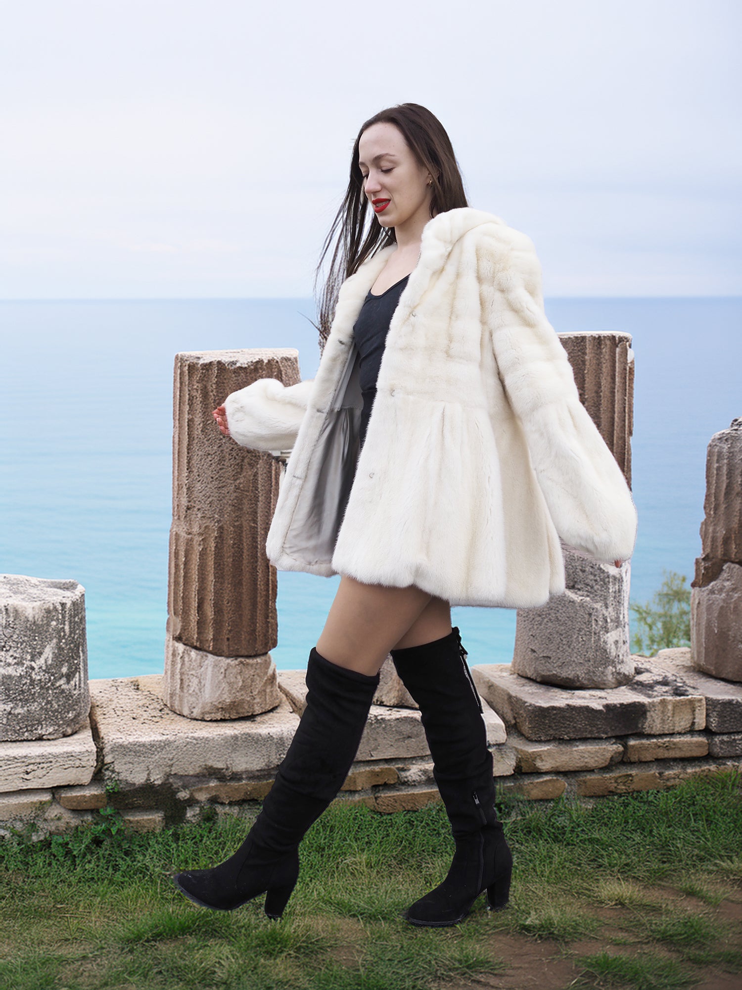 Elegant Beige Pearl Mink Fur Coat with hood, worn by a model near ancient ruins overlooking the sea. Luxurious warmth with hook-eye closure and satin lining.
