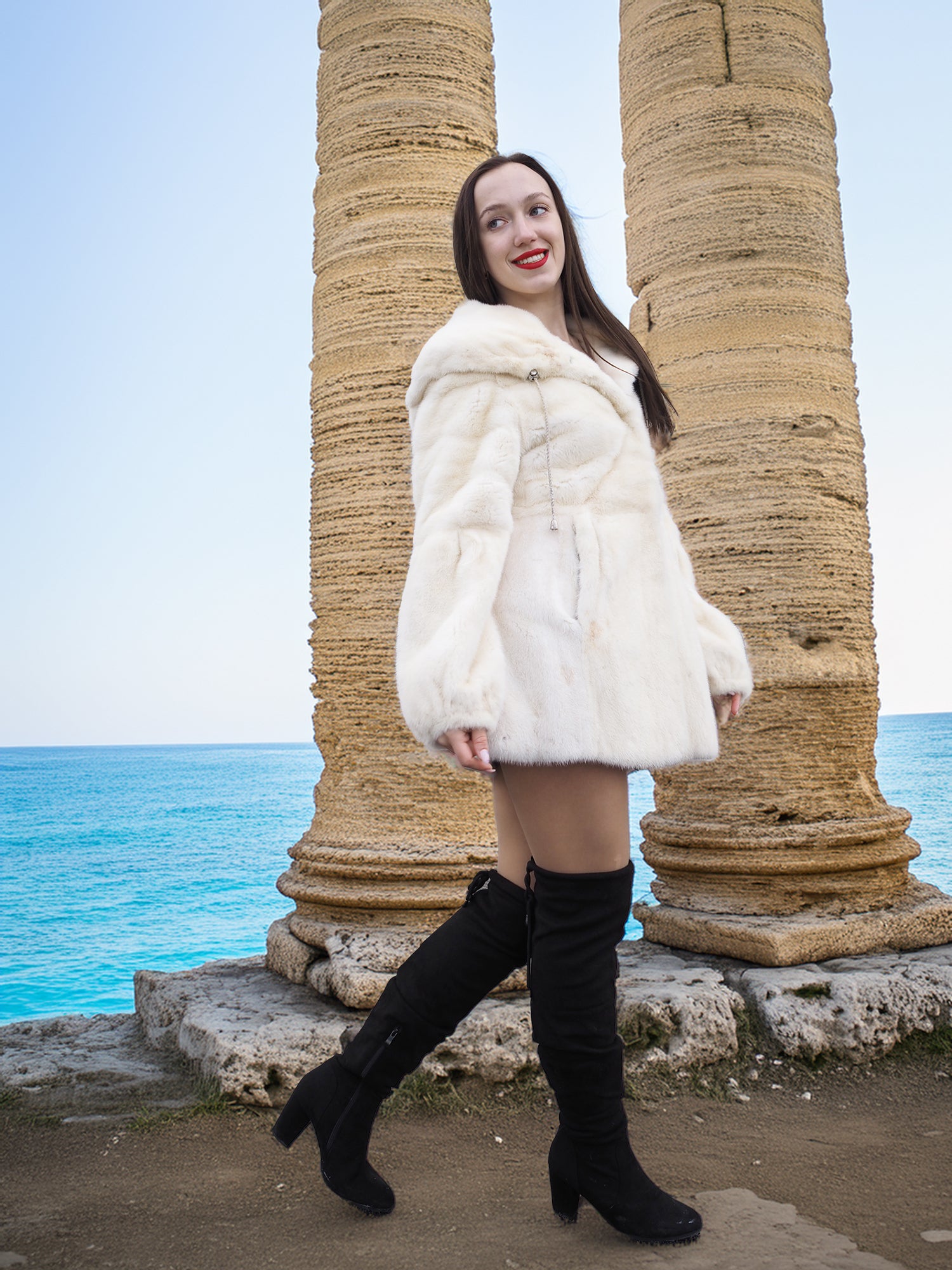 Elegant woman wearing a Beige Pearl Mink Fur Coat with a hood, standing by ancient stone columns near the sea. Chic warmth with hook-eye closure.