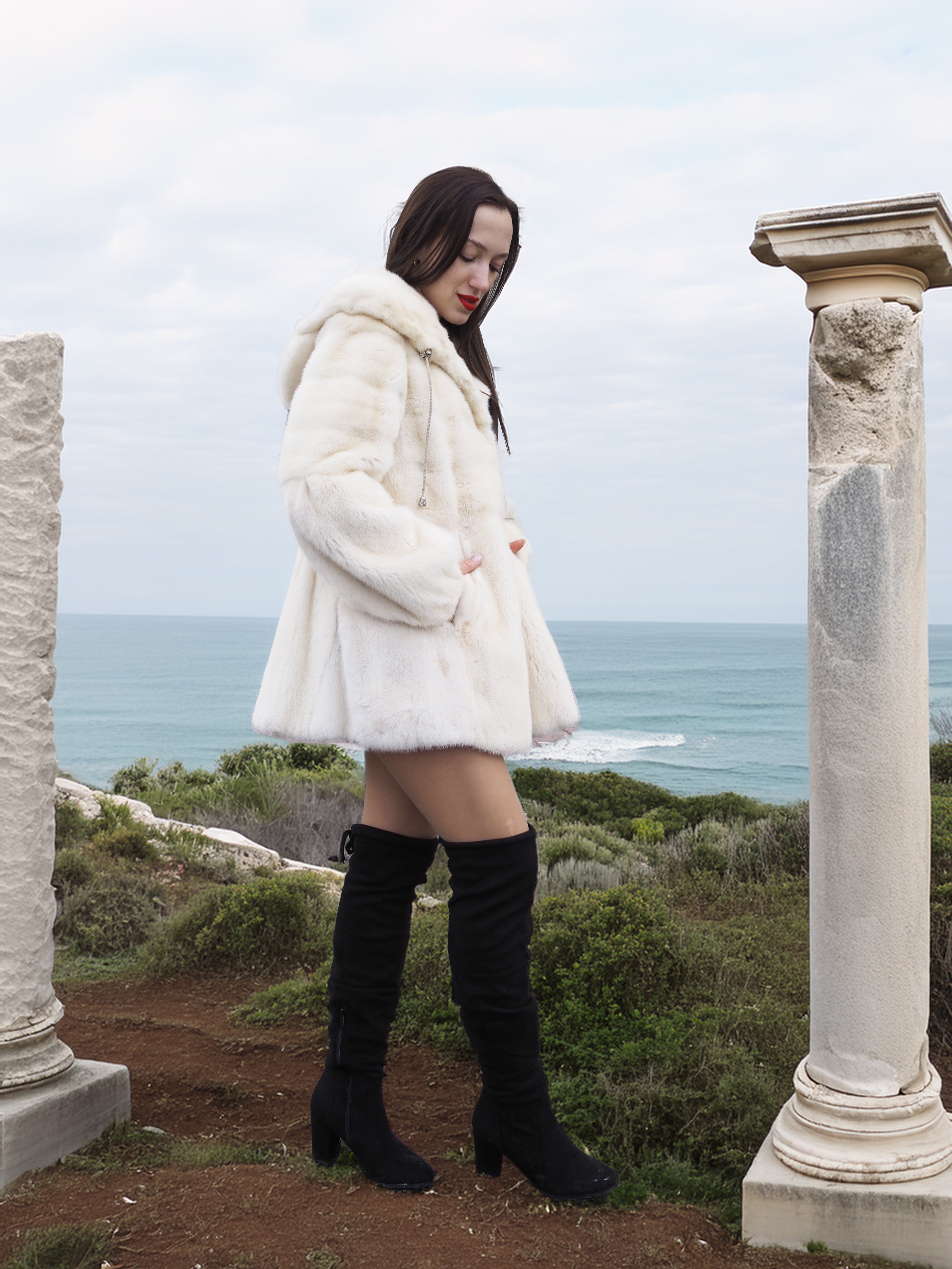 Elegant woman wearing a Beige Pearl Mink Fur Coat with a hood, standing between ancient columns overlooking the ocean. Chic warmth with hook-eye closure.