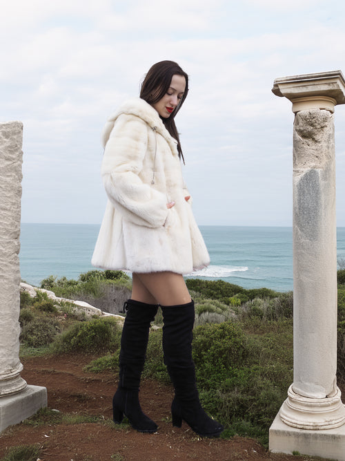 Elegant woman wearing a Beige Pearl Mink Fur Coat with a hood, standing between ancient columns overlooking the ocean. Chic warmth with hook-eye closure.
