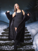 Woman in a long black fur coat standing on snow-covered steps with a dark, forested background.