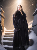 Woman in a long black coat standing on snow-covered steps with a warm light source in the background