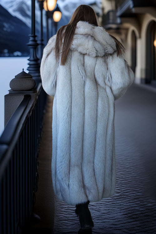 Person wearing a long white fur coat standing on a snowy path with mountains in the background.