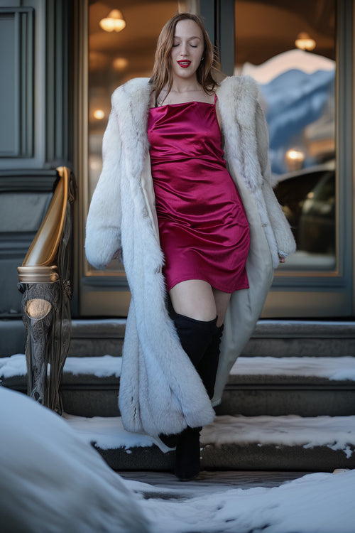 Woman in a pink dress and white fur coat standing on snowy steps with mountains in the background