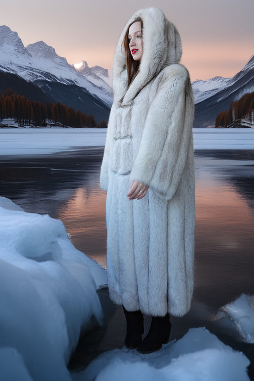 Person wearing a long fur coat standing in a snowy landscape with mountains and a lake.
