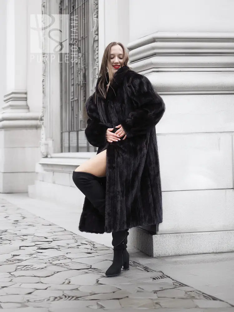 Black mink fur coat with long length, notched collar, and side pockets; model stands in profile.