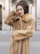 Beige mink fur coat with pointed collar and button-front, hands clasped.