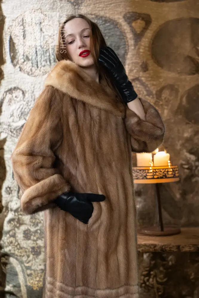 Tan mink fur hooded coat with ribbed hem detail; black gloves in a warm indoor setting
