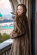 Women’s dark brown mink coat with hood and voluminous sleeves, side view on balcony.