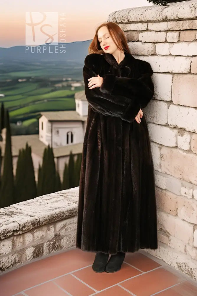 Black mink coat in long cut with high collar, model leaning on stone wall.