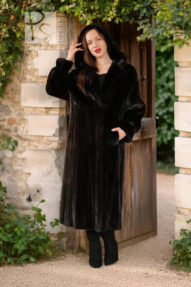 Black hooded mink coat with full sleeves and long skirt; model posing with hand near hair.