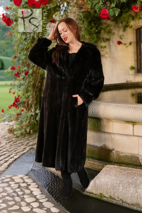 Black hooded mink coat with long sleeves and open front; model standing on cobblestones.
