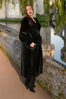 Black hooded mink coat with relaxed fit and long hem; model resting hand on coat collar.