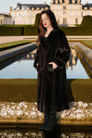 Black hooded mink coat, long and straight; model standing by a reflecting pool.