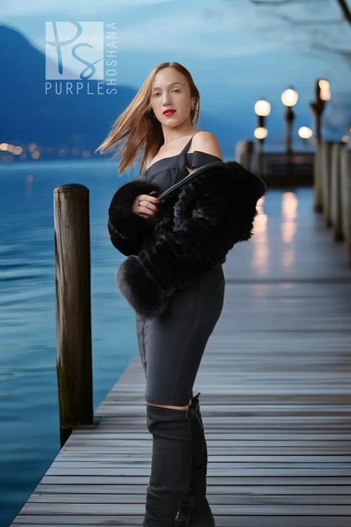 A woman stands on a wooden dock by the water at dusk, wearing a black off-the-shoulder top, dark gray fitted pants, black thigh-high boots, and holding a black faux-fur accessory. Mountains and lake are visible in the background.
