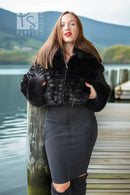 A woman stands on a wooden dock by a lake wearing a black cropped faux-fur jacket and a dark gray fitted skirt, with mountains in the background.