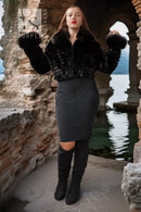 A woman stands in a stone archway by the water wearing a black faux-fur cropped jacket with fluffy cuffs, a dark gray fitted pencil skirt, and black over-the-knee boots.