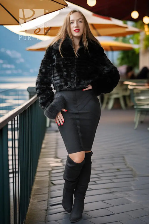 A woman stands on an outdoor patio by a railing, wearing a black faux-fur cropped jacket, a fitted black mini skirt with a front cutout, and black over-the-knee boots.