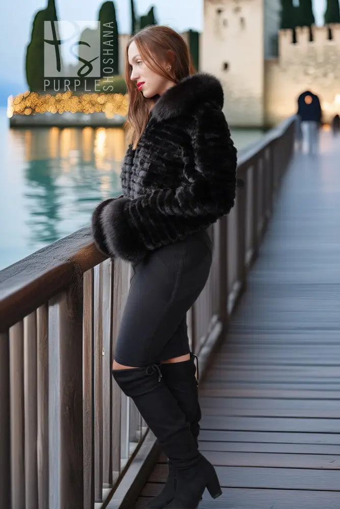 A woman stands on a wooden boardwalk by the water wearing a black faux fur cropped jacket, black leggings, and black over-the-knee boots, with a castle and water in the background.