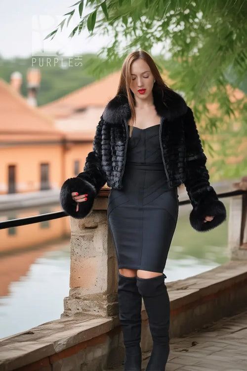 A woman stands outdoors by a stone railing next to a canal, wearing a black fur-trimmed cropped jacket over a black strapless dress and black over-the-knee boots.
