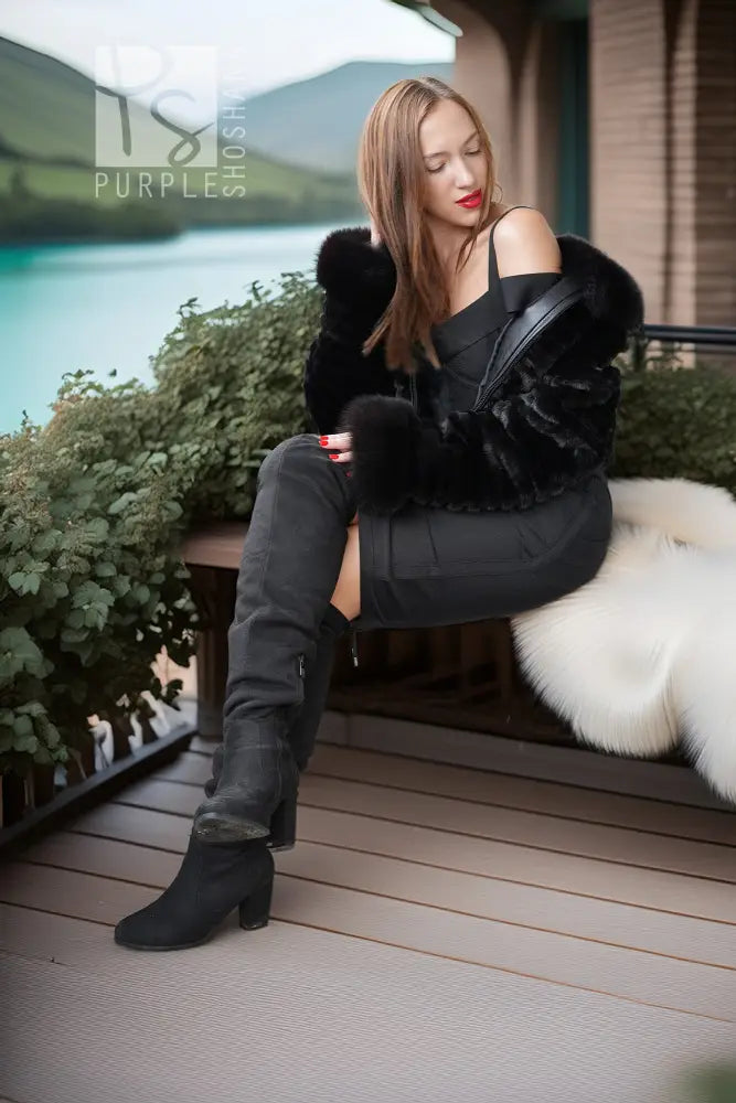 A woman sits on a wooden bench on a deck, wearing a black off-the-shoulder dress, a black faux-fur jacket, black over-the-knee boots, and holding a black shoulder bag. A scenic lake and mountains are visible in the background.