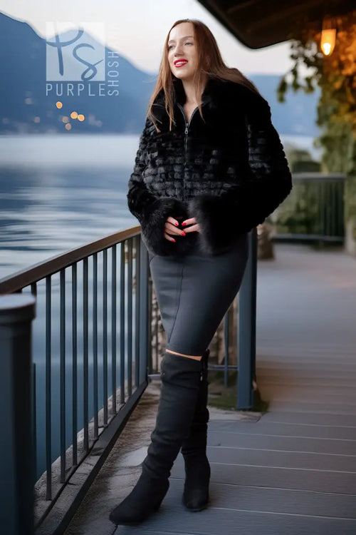 A woman stands on a lakeside boardwalk wearing a black faux-fur cropped jacket, a dark gray fitted skirt, and black over-the-knee boots, with mountains and water in the background.