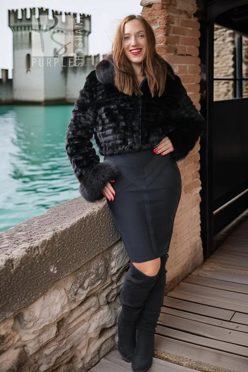 A woman posing outdoors in a black faux fur cropped jacket, a black bodycon skirt, and black over-the-knee boots, with a stone wall, brick wall, and water in the background.
