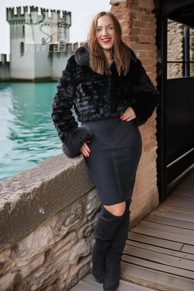 A woman posing outdoors in a black faux fur cropped jacket, a black bodycon skirt, and black over-the-knee boots, with a stone wall, brick wall, and water in the background.