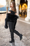 A woman in a black off-the-shoulder top, black pants, and black over-the-knee boots stands on a cobblestone walkway in an outdoor architectural setting, holding a black faux-fur wrap around her arms.