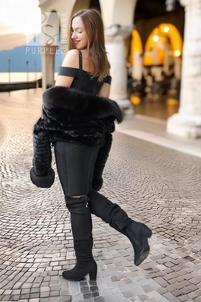 A woman in a black off-the-shoulder top, black pants, and black over-the-knee boots stands on a cobblestone walkway in an outdoor architectural setting, holding a black faux-fur wrap around her arms.