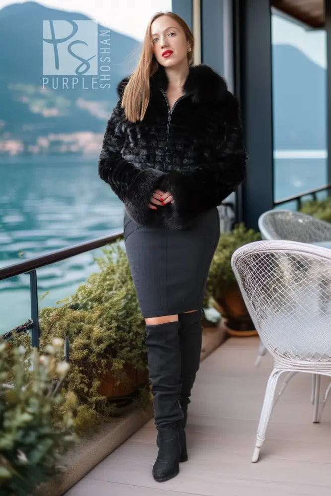 A woman stands on a balcony overlooking water and mountains, wearing a black faux-fur cropped jacket, a dark gray fitted midi skirt with a thigh cutout, and black over-the-knee boots.