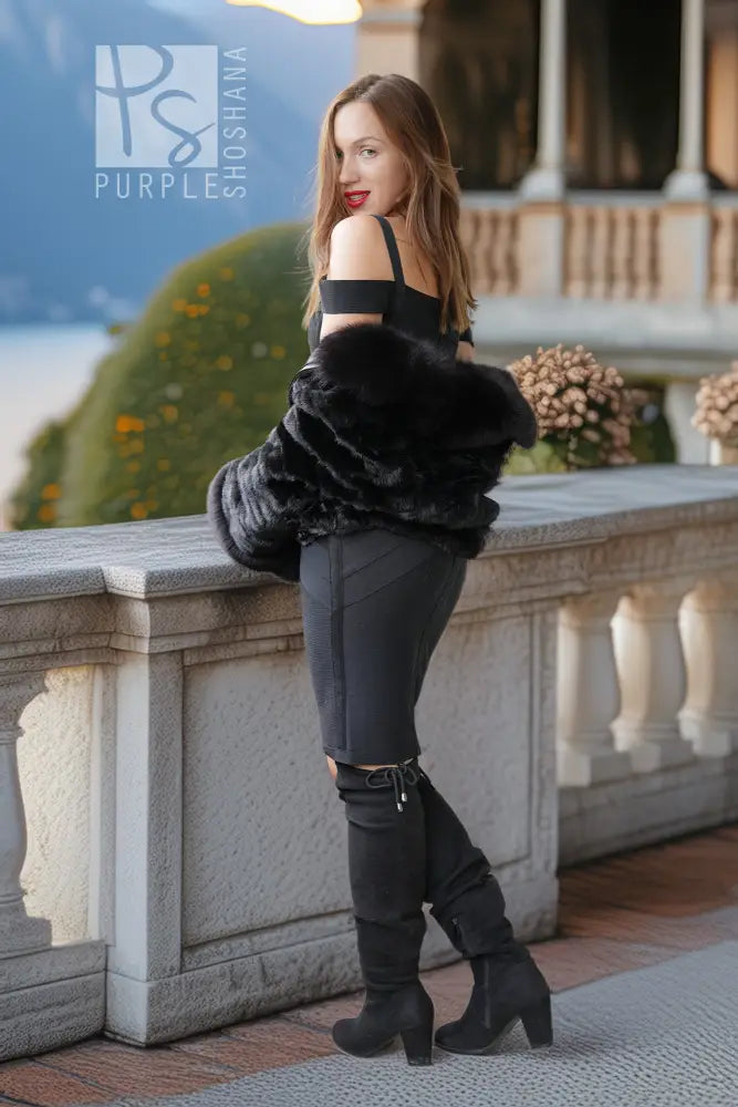 A woman stands on an outdoor terrace wearing a black off-the-shoulder top, black pants, black thigh-high boots, and holding a black faux-fur jacket. The background features a stone balustrade, greenery, and a distant mountain landscape.