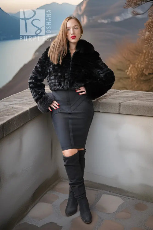 A woman posing on a stone balcony overlooking a scenic lake and mountains, wearing a black faux fur cropped jacket, a black bodycon pencil skirt with a thigh cutout, and black over-the-knee boots.