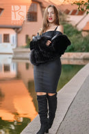 A woman stands outdoors beside a canal at sunset, wearing a black off-the-shoulder mini dress, a voluminous black faux-fur stole, and black over-the-knee boots.