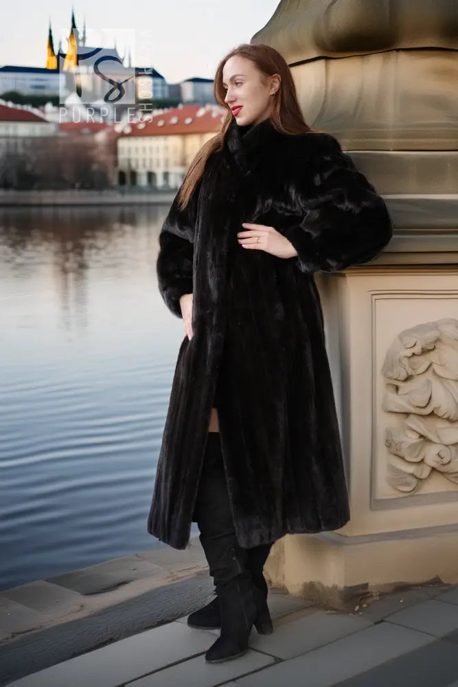 Black mink coat with rounded neckline and voluminous sleeves, standing by a riverside wall.