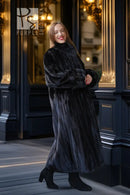 Black mink fur long coat with high collar and button front, portrait view beside dark wall.