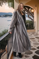 A woman wearing a long, luxurious gray fur coat over a black dress and black boots, standing on a stone walkway of a coastal terrace at sunset.